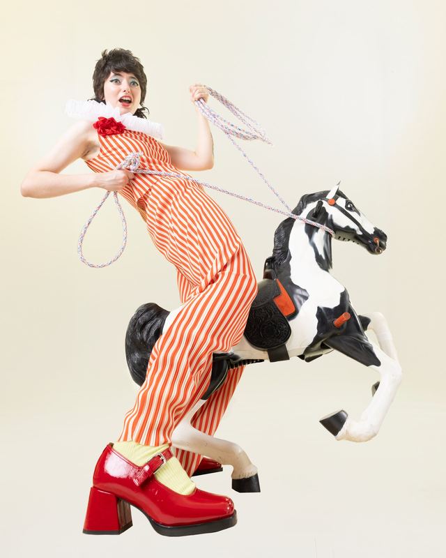 Riding a carousel horse in a striped dress, Sarah Squirm by David Spector